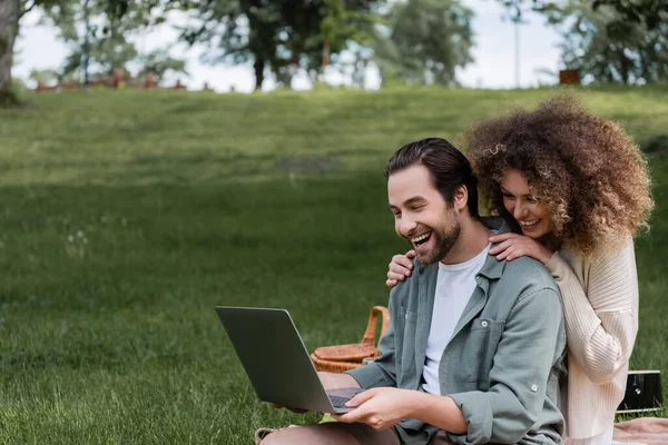 Uomo stupito utilizzando laptop vicino donna felice durante il picnic nel parco — Foto stock