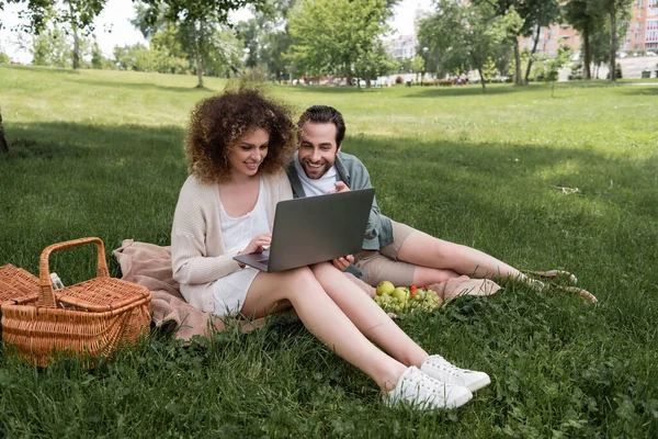 Happy woman using laptop while sitting on blanket with boyfriend during picnic — Fotografia de Stock