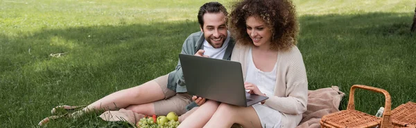 Щаслива жінка використовує ноутбук, сидячи на ковдрі з хлопцем під час пікніка, банер — стокове фото