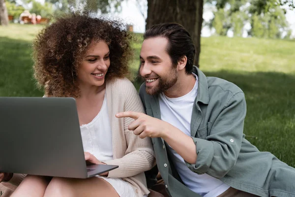 Glücklicher Mann zeigt mit Finger auf Laptop neben fröhlicher Frau — Stockfoto