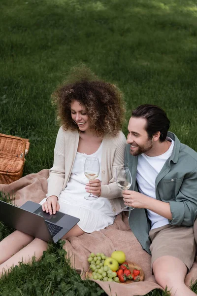 Высокий угол обзора пары, держащей бокал вина и глядящей на ноутбук во время пикника — стоковое фото