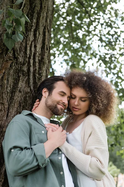 Joven y feliz pareja con los ojos cerrados abrazando cerca del tronco del árbol - foto de stock