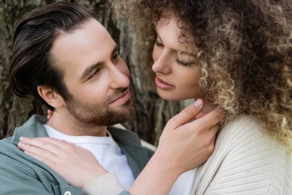 Portrait of young and happy man touching face of curly woman near tree trunk — Fotografia de Stock