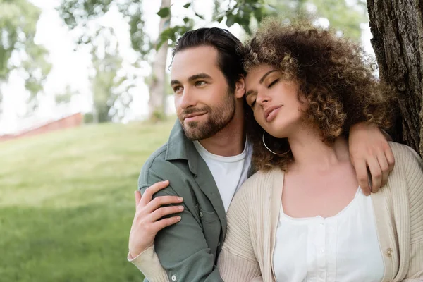 Glücklicher Mann umarmt lockige junge Frau mit geschlossenen Augen in der Nähe des Baumstammes — Stockfoto