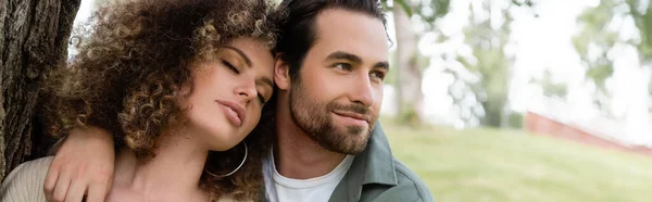 Happy man embracing curly and young woman with closed eyes near tree trunk, banner — Photo de stock