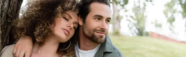 Hombre feliz abrazando rizado y mujer joven con los ojos cerrados cerca del tronco del árbol, pancarta - foto de stock