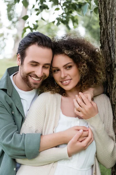 Happy man embracing curly and positive woman near tree trunk — Stockfoto