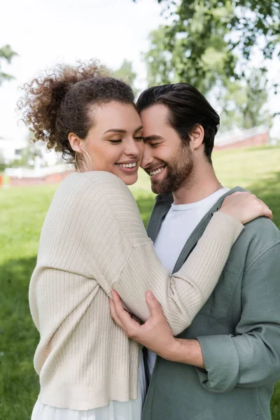Homem barbudo alegre abraçando com mulher encaracolada e alegre no parque de verão verde — Fotografia de Stock