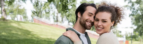 Homme barbu heureux étreignant avec une femme bouclée et joyeuse dans un parc d'été vert, bannière — Photo de stock