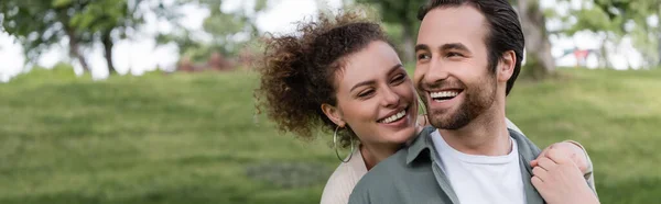 Pleased and curly woman hugging boyfriend from behind in green summer park, banner — Photo de stock