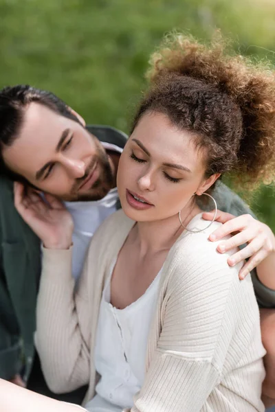 Tender and curly woman touching face of bearded boyfriend in park - foto de stock