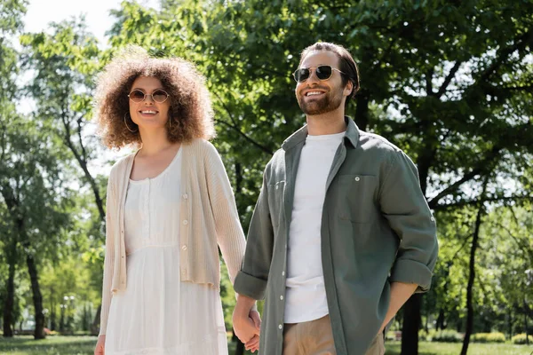 Stylisches und glückliches Paar mit Sonnenbrille hält Händchen im Park — Stockfoto