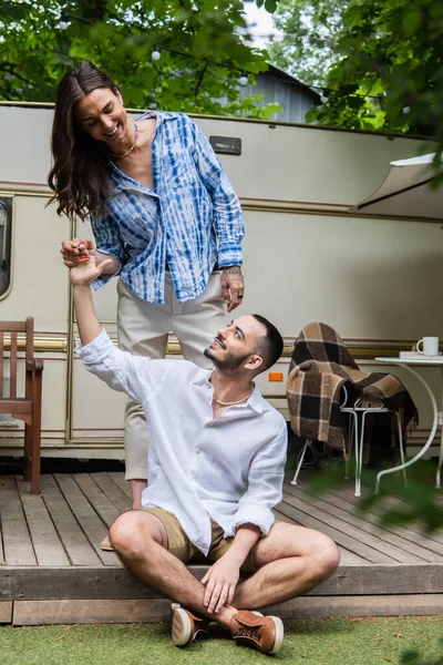 Cheerful tattooed gay man holding hand of bearded boyfriend near travel van - foto de stock