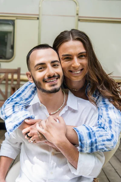 Portrait of happy tattooed gay man hugging cheerful bearded boyfriend near travel van - foto de stock