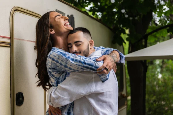 Happy gay man with tattoo smiling while hugging boyfriend near van — Stock Photo