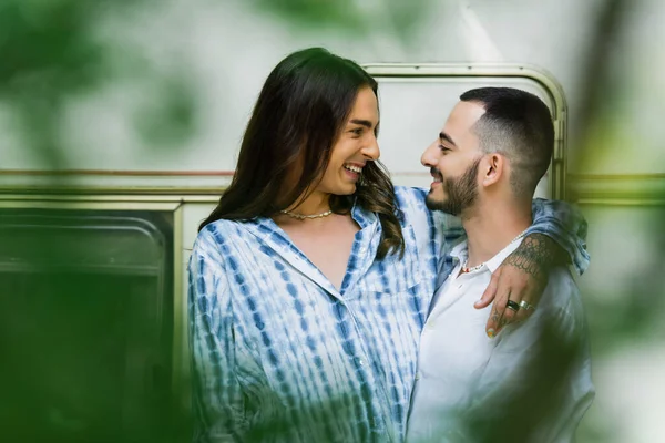 Glückliches homosexuelles Paar lächelt, während es sich in der Nähe eines Lieferwagens mit verschwommenen Blättern im Vordergrund umarmt — Stockfoto