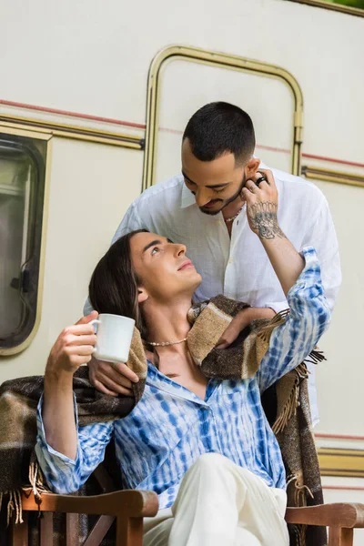 Caring gay man holding blanket near tattooed boyfriend with cup of coffee sitting near van - foto de stock