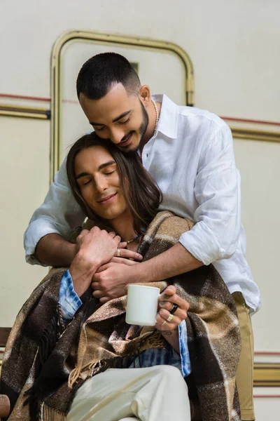 Bearded gay man hugging boyfriend in blanket sitting with cup of coffee near van — Photo de stock