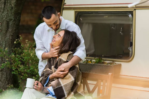 Caring gay man hugging cheerful boyfriend in blanket sitting with cup near van — Stock Photo