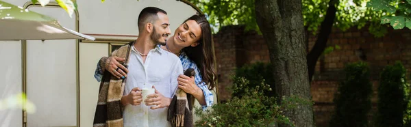 Happy tattooed man hugging bearded boyfriend in blanket standing with cup near van, banner — Stock Photo