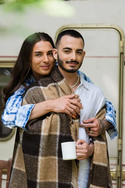 Smiling man with tattoo hugging bearded boyfriend in blanket near blurred van — Stock Photo