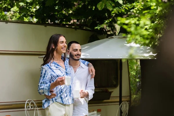 Feliz pareja gay sosteniendo tazas de café mientras está de pie cerca de van durante el viaje en verano - foto de stock