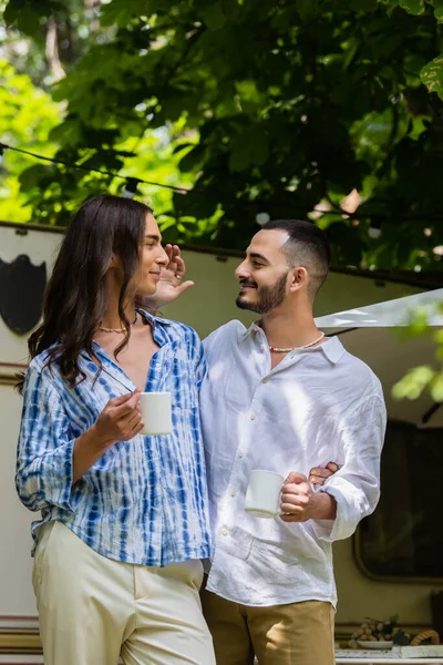 Happy gay couple holding cups of coffee while looking at each other near van during journey in summer — Stock Photo