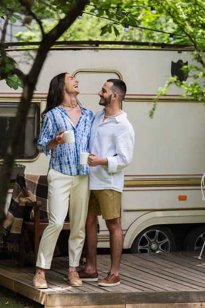 Full length of happy gay couple holding cups of coffee while laughing near van in summer — Fotografia de Stock