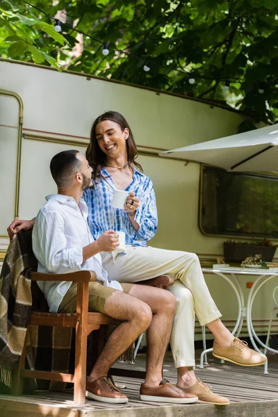 Happy gay man holding cup of coffee while sitting with boyfriend near van during journey in summer — Stockfoto