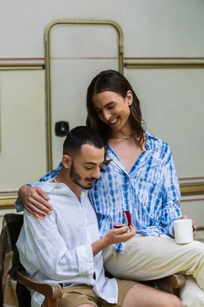 Gay man holding jewelry box with ring near happy boyfriend with cup of coffee — Fotografia de Stock