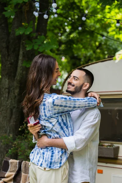 Gay man holding jewelry box with wedding ring and hugging happy boyfriend during journey in summer — Photo de stock