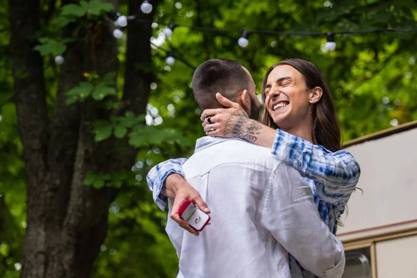 Tattooed gay man holding jewelry box with wedding ring and hugging boyfriend in summer — Photo de stock
