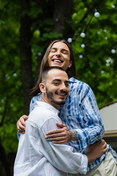 Glückliches homosexuelles Ehepaar lächelt, während es sich in der Nähe eines verschwommenen Reisebusses umarmt — Stockfoto