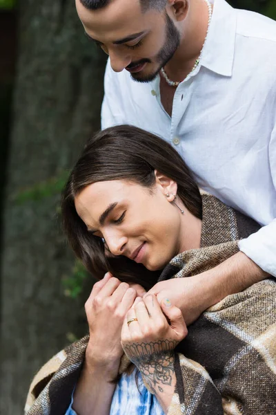 Bearded gay man hugging smiling husband in blanket with wedding ring on finger - foto de stock