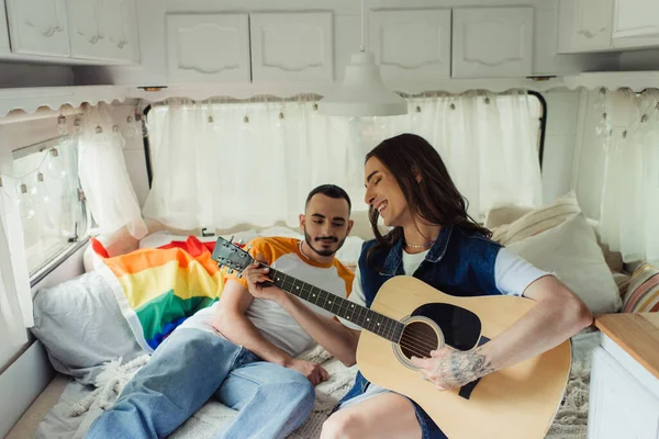 Happy and tattooed gay man playing acoustic guitar near boyfriend on bed in modern van - foto de stock