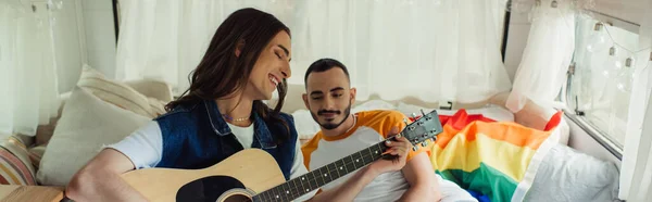 Happy and tattooed gay man playing acoustic guitar near boyfriend on bed in modern van, banner - foto de stock