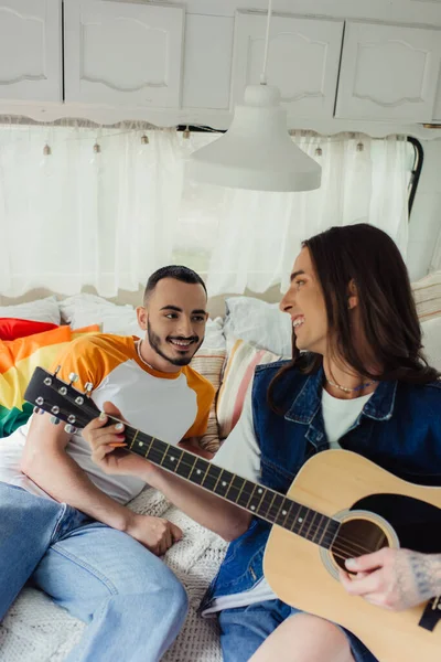 Feliz y tatuado gay hombre jugando acústica guitarra cerca novio y lgbt bandera en cama en van - foto de stock