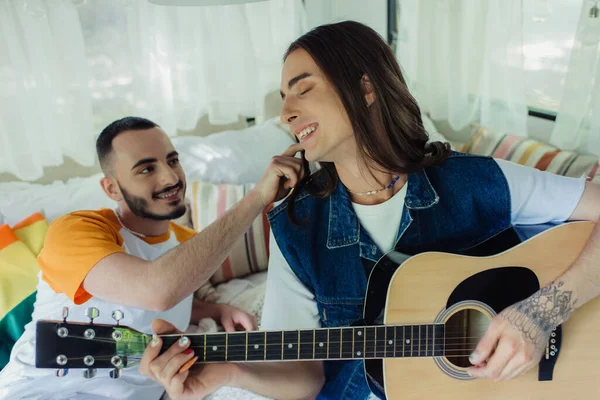 Cheerful and tattooed gay man playing acoustic guitar near boyfriend on bed in modern van — Foto stock