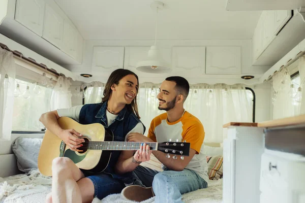 Happy gay man playing acoustic guitar near bearded boyfriend on bed in modern van - foto de stock