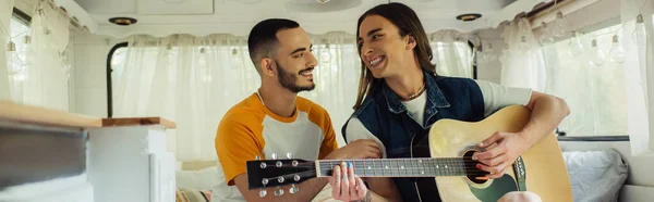 Happy and tattooed gay man playing acoustic guitar near boyfriend in modern van, banner — Stockfoto