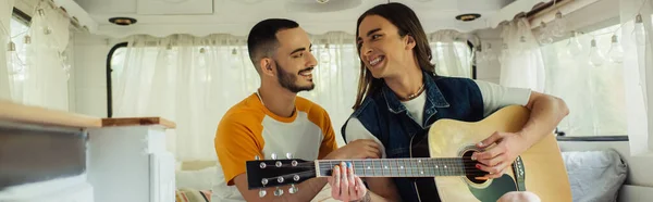 Happy and tattooed gay man playing acoustic guitar near boyfriend in modern van, banner - foto de stock