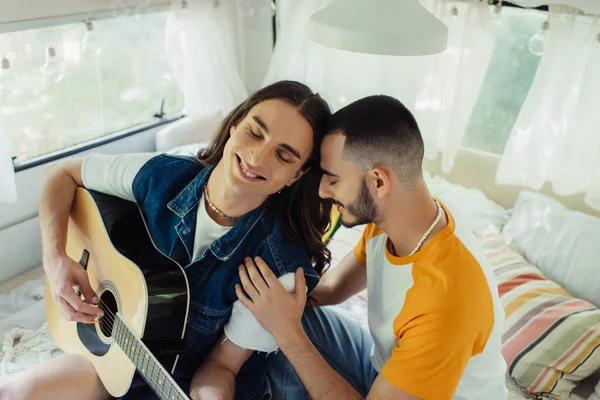 High angle view of happy gay man with long hair playing acoustic guitar near boyfriend on bed in modern van - foto de stock