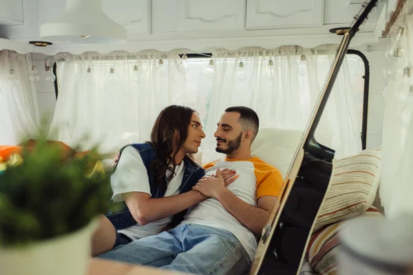 Gay man with long hair hugging bearded boyfriend on bed near guitar in modern van - foto de stock