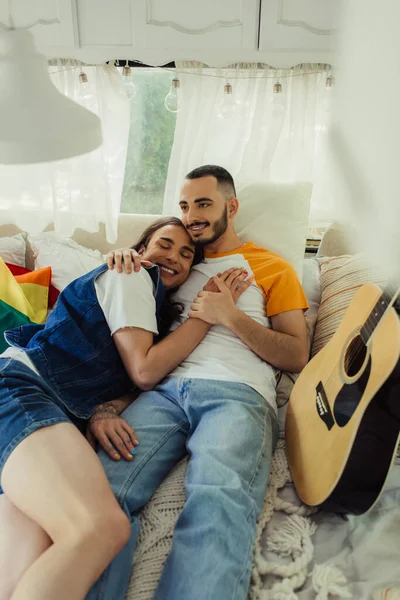 Smiling gay man with tattoo lying in bed with bearded boyfriend near lgbt flag in modern van — Foto stock