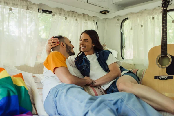 Sorridente gay uomo con tatuaggio guardando barbuto fidanzato su letto vicino lgbt bandiera e acustica chitarra in moderno van — Foto stock