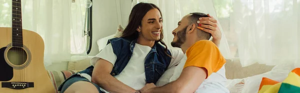 Uomo gay sorridente con tatuaggio guardando il fidanzato barbuto sul letto vicino alla bandiera lgbt e chitarra acustica nel furgone, banner — Foto stock