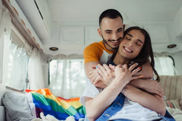 Smiling gay man hugging pleased boyfriend with lgbt flag on blurred background in modern van — Photo de stock