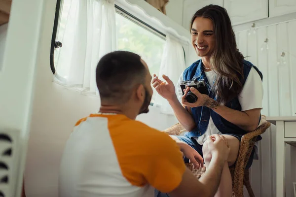 Blick auf einen glücklichen homosexuellen Mann, der eine Vintage-Kamera hält und in einem Sessel in der Nähe seines Freundes in einem Van sitzt — Stockfoto