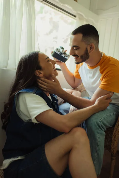 Bearded gay man holding vintage camera while sitting in armchair near happy boyfriend in van — Stock Photo