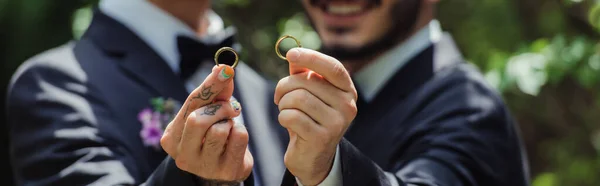 Cropped view of happy and blurred gay men holding wedding rings in hands, banner — стоковое фото